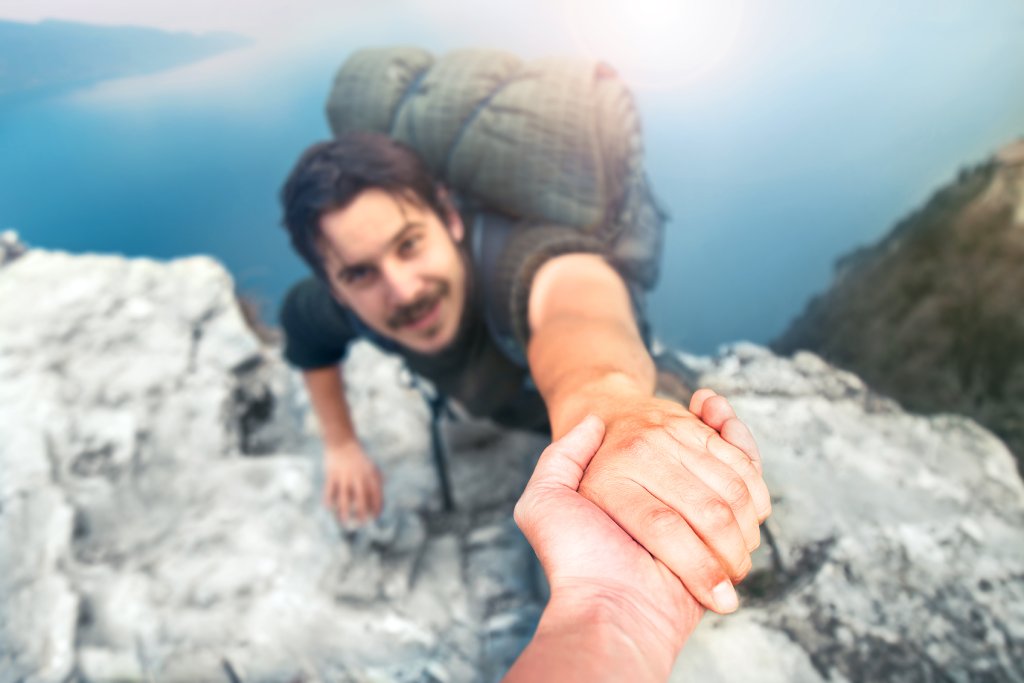 A hiker on a rock ledge gripping an outstretched hand that is pulling him up